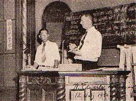 Old sepia photograph of W. Edwards Deming lecturing at a desk in Tokyo, a man standing beside him.