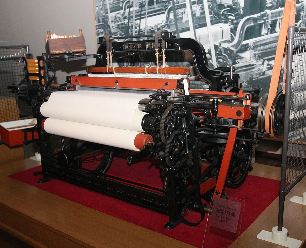 A Toyoda Type G automatic loom on display in a museum: black iron frame, white cloth rolls and orange belts.