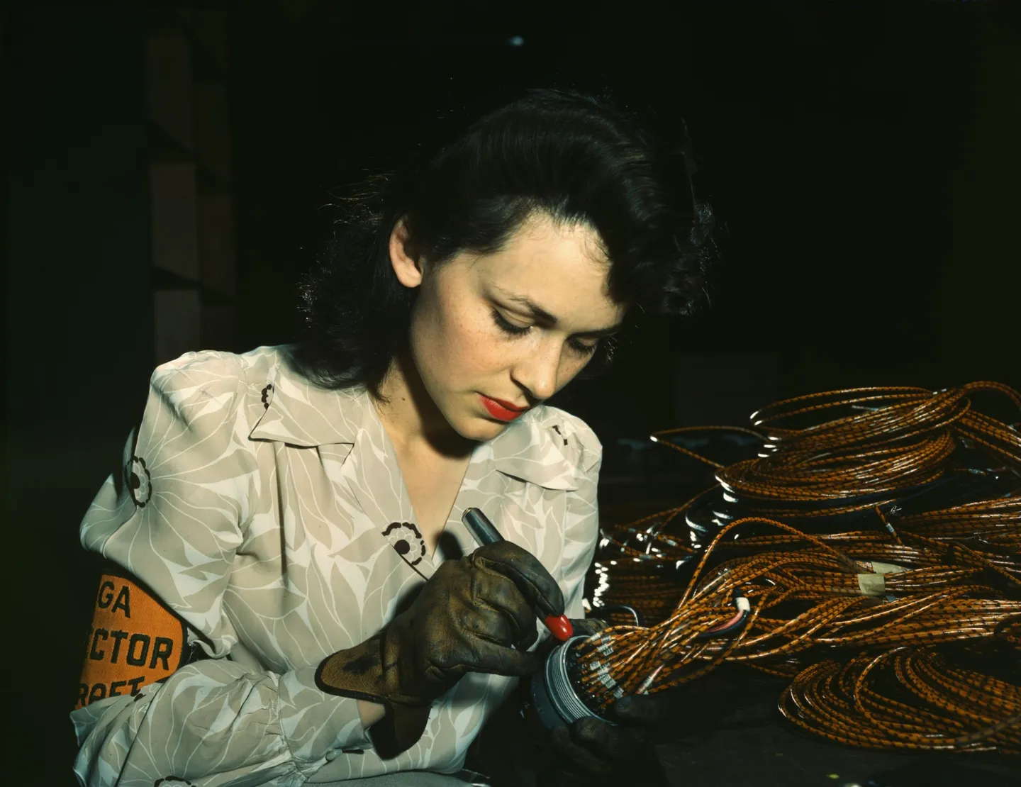 Colour photograph of a young woman in work gloves and an inspector's armband, checking the end of a bundle of aircraft wiring.